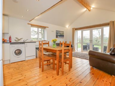 A dining area with wooden table and chairs next to a kitchen with washing machine and sink in an open living space at Vine Cottage in St Ewe near Sticker