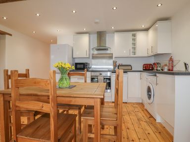 A kitchen with wooden dining table and chairs a vase with yellow flowers white cabinets and appliances at Vine Cottage St Ewe near Sticker 