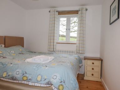 A bedroom with a bed covered in a floral duvet beside a window with curtains and a bedside table at Vine Cottage in St Ewe near Sticker 