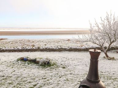 A snow-covered garden with a chiminea tree and stone wall near a body of water at Sonya's Cottage Anthorn Solway Firth