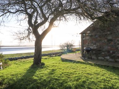 A backyard with a tree that has a swing hanging from it a stone patio with a barbecue and chairs next to a stone house near a body of water at Sonya's Cottage Anthorn Solway Firth