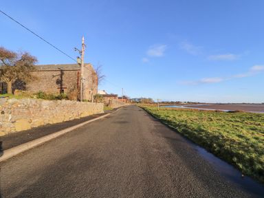 A paved road next to stone buildings and grassy area by water at Sonya's Cottage in Anthorn Solway Firth