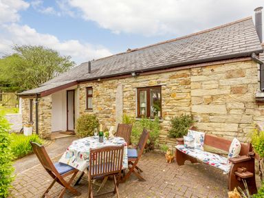 A stone cottage exterior with a table and four chairs and a wooden bench with cushions at Poldark Cottage in East Taphouse