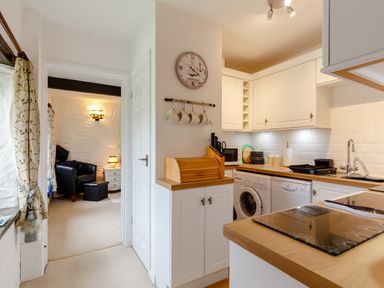 A kitchen with white cabinets wooden countertops and appliances next to a sitting room with black armchairs at Poldark Cottage East Taphouse