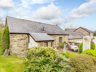 A stone cottage with a slate roof surrounded by bushes and a patio with a table and chairs at Poldark Cottage in East Taphouse