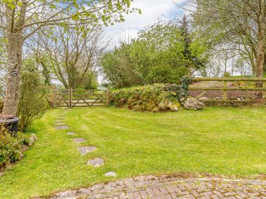 A garden with a grassy lawn stone path trees and wooden gates at Poldark Cottage in East Taphouse