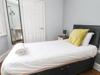 A bedroom with a single bed with white sheets and a yellow pillow beside a nightstand with a lamp and a door with a mirror in Mountain View in Penmaenmawr
