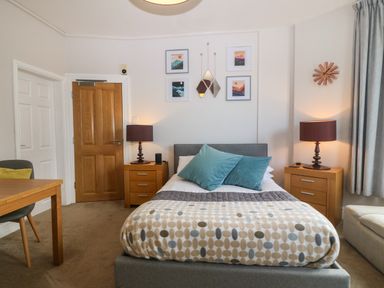 A bedroom with a bed with pillows and a patterned blanket two wooden nightstands with lamps a wooden table with a chair wall art and a clock at Y Castell Apartment 1 in Criccieth