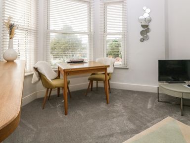 A room with a wooden table and two chairs with white throws near windows with blinds a round table with a television and a wall decoration with round mirrors at Y Castell Apartment 2 in Criccieth
