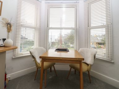 A small dining area with a wooden table and two chairs next to three windows with blinds at Y Castell Apartment 2 in Criccieth