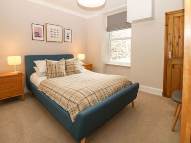 A bedroom with a blue upholstered bed beige bedding two bedside tables with lamps and framed pictures on the wall at Y Castell Apartment 2 in Criccieth