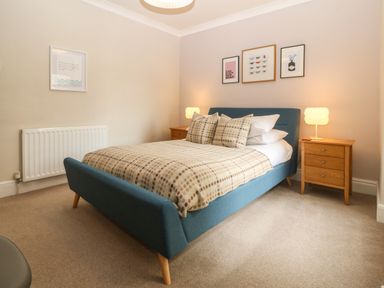 A bedroom with a blue upholstered bed a blanket and cushions two wooden bedside tables with lamps and framed pictures on the walls at Y Castell Apartment 2 in Criccieth