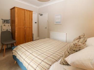 A bedroom with a bed covered by a patterned blanket pillows a wooden wardrobe a gray chair and an ironing board at Y Castell Apartment 2 in Criccieth