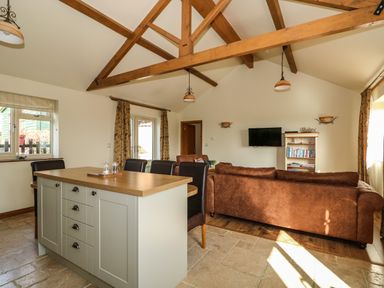 A living room with brown sofas a mounted TV a bookshelf and a kitchen island with chairs at Sunnyside Cottage in Blackborough End near Kings Lynn