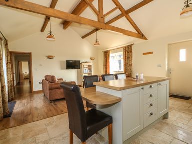 An open-plan kitchen and living area with wooden beams a kitchen island leather chairs and a wall-mounted television at Sunnyside Cottage in Blackborough End near King's Lynn