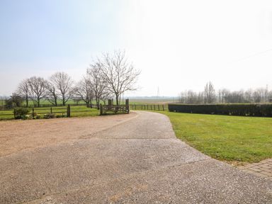 A paved driveway leading to a wooden gate with grassy areas and trees in the background at Sunnyside Cottage in Blackborough End near King's Lynn