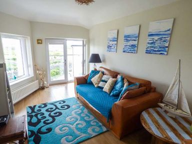 A living room with a brown sofa with blue cushions and a patterned rug and a small wooden table with a model sailboat at Ty'r Enfys Bach - Upper in Trearddur Bay