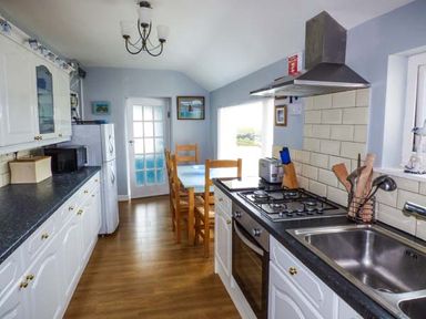A kitchen with white cabinets black countertops a gas stove wooden dining table and chairs at Ty'r Enfys Bach - Upper in Trearddur Bay
