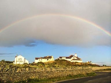 A rainbow over houses on a hill with a stone wall and road at Ty'r Enfys Bach - Upper in Trearddur Bay