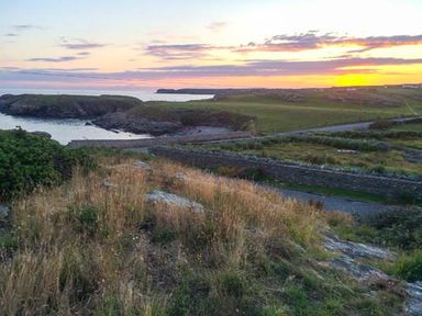 A coastal landscape with grassy fields stone walls and a sunset over the sea at Ty'r Enfys Bach - Upper in Trearddur Bay