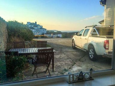 A patio with a wooden table and chairs next to a white pickup truck and a view of houses and the sea at Ty'r Enfys Bach - Upper in Trearddur Bay