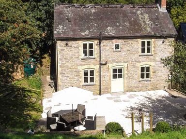 A stone house with four windows and a door beside a patio with outdoor furniture and an umbrella at Ploony Cottage in Bleddfa