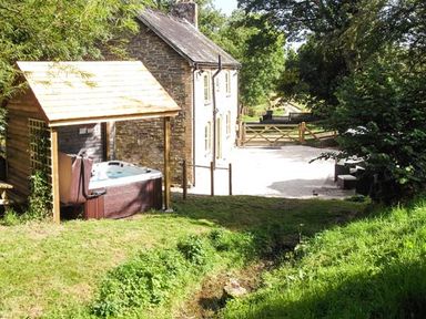 An outdoor area with a wooden structure covering a hot tub a stone house a grass lawn and a gravel driveway with a wooden gate at Ploony Cottage in Bleddfa