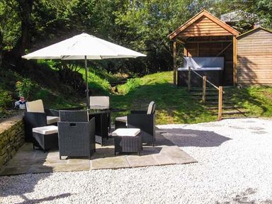 An outdoor seating area with chairs and a table under an umbrella and a wooden gazebo with a hot tub in a garden at Ploony Cottage in Bleddfa