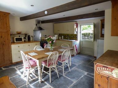 A kitchen with a wooden table and six chairs with cushions a vase of flowers on the table cupboards a microwave a stove and a door leading outside at Ploony Cottage in Bleddfa