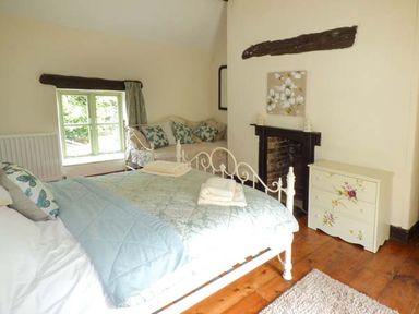 A bedroom with a bed covered with light blue bedding a small couch by a window a fireplace and a painted chest of drawers at Ploony Cottage in Bleddfa