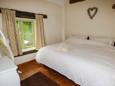 A bedroom with a large bed white bedding wooden floor small window with curtains and a heart decoration on wall at Ploony Cottage in Bleddfa