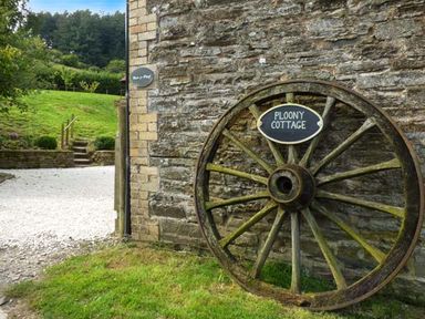 A large wooden wagon wheel leaning against a stone wall with a sign that reads Ploony Cottage in a grassy outdoor area at Ploony Cottage in Bleddfa