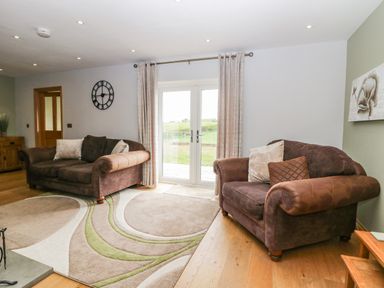 A living room with two brown sofas and patterned rug near glass doors with curtains at Primrose Cottage in Aldingham