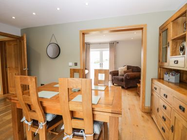 A dining room with wooden table and chairs next to a wooden cabinet and an adjoining room with armchair and glass doors at Primrose Cottage in Aldingham