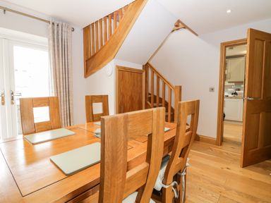 A dining area with a wooden table and chairs near wooden stairs and a doorway to a kitchen at Primrose Cottage in Aldingham