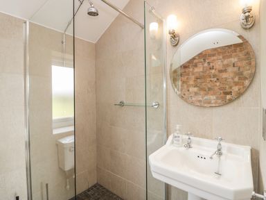 A bathroom with a glass enclosed shower a white sink an oval mirror and a tiled wall at Primrose Cottage in Aldingham
