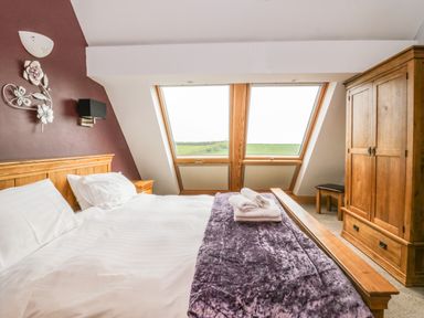 A bedroom with a wooden bed, white bedding, purple throw, two folded towels, wooden wardrobe, and two large windows at Primrose Cottage in Aldingham