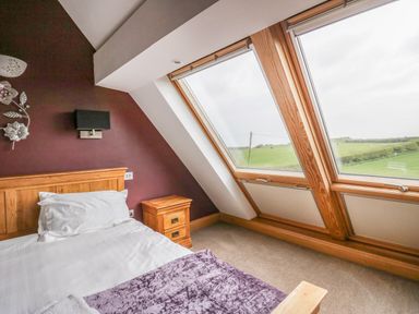 A bedroom with a bed a wooden nightstand and slanted windows showing a green field at Primrose Cottage in Aldingham