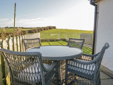 An outdoor patio with a round glass table and four wicker chairs overlooking a grassy field at Daisy Cottage in Aldingham