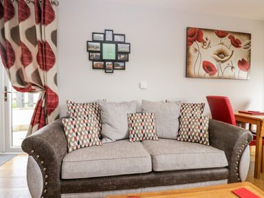A living room with a sofa with patterned cushions a floral painting on the wall and red and beige curtains at Daisy Cottage in Aldingham