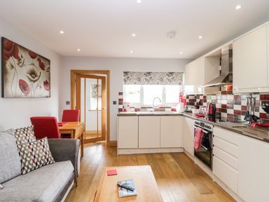 A kitchen with white cabinets red and white tiled backsplash a wooden floor a gray sofa with patterned cushions and a wooden table with red chairs at Daisy Cottage in Aldingham