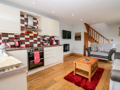 An open kitchen and living area with cream cabinets red and brown tiled backsplash a wooden coffee table on a red rug and a grey sofa at Daisy Cottage in Aldingham