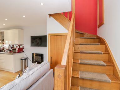 A wooden staircase with carpet runners next to a living area with a grey sofa and a kitchen with white cabinets and red appliances at Daisy Cottage in Aldingham