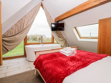 A bedroom with a red blanket on the bed a bathtub near a large triangular window and a wall-mounted television at Daisy Cottage in Aldingham