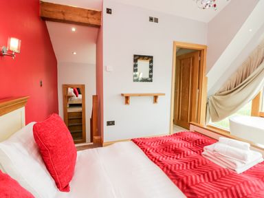 A bedroom with a bed with red pillows and a red blanket with towels placed on it a wall with a small shelf and a mirror and a door leading to another room at Daisy Cottage in Aldingham