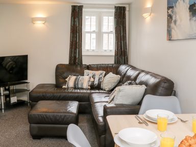 A living room with a brown leather sectional sofa with pillows, a matching ottoman, a television on a stand, and a dining table set with plates and glasses of orange juice at Razorbill Cottage in Whitby