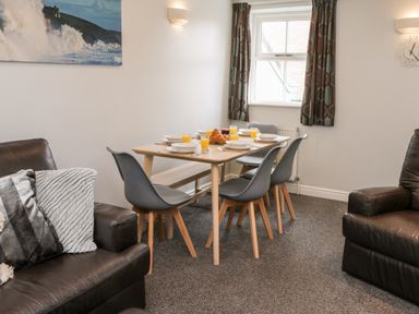 A dining area with a wooden table set with plates glasses of orange juice and croissants surrounded by grey chairs and brown leather sofas at Razorbill Cottage in Whitby