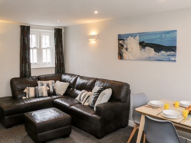 A living room with a black leather sectional sofa and ottoman with cushions next to a dining table set with plates and glasses at Razorbill Cottage in Whitby