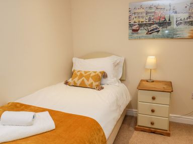 A single bed with white bedding and a decorative pillow next to a wooden nightstand with a lamp and a framed artwork above at Razorbill Cottage in Whitby