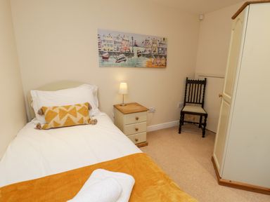 A small bedroom with a single bed with a yellow cushion and blanket beside a bedside table with a lamp a wooden chair and a wardrobe at Razorbill Cottage in Whitby